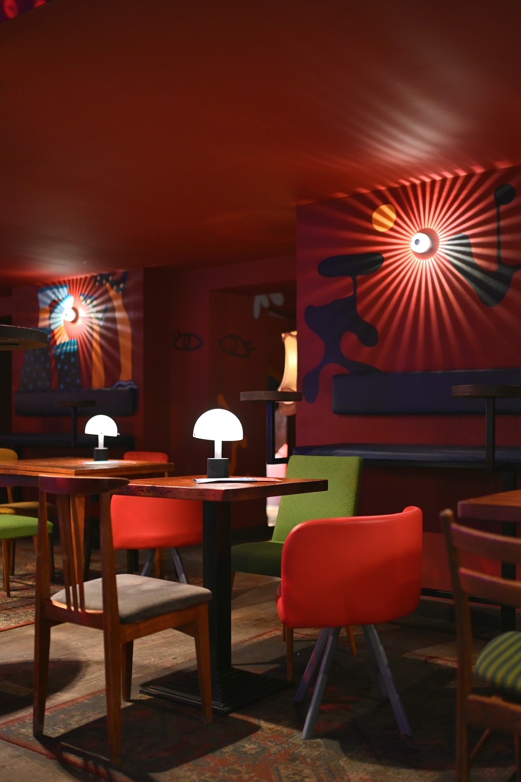 Cozy seating area with a mustard yellow vintage couch, green chair, blue and orange round ottomans, a wooden coffee table, a lit floor lamp, and an abstract mural on a dark blue wall reflected in a corner mirror.