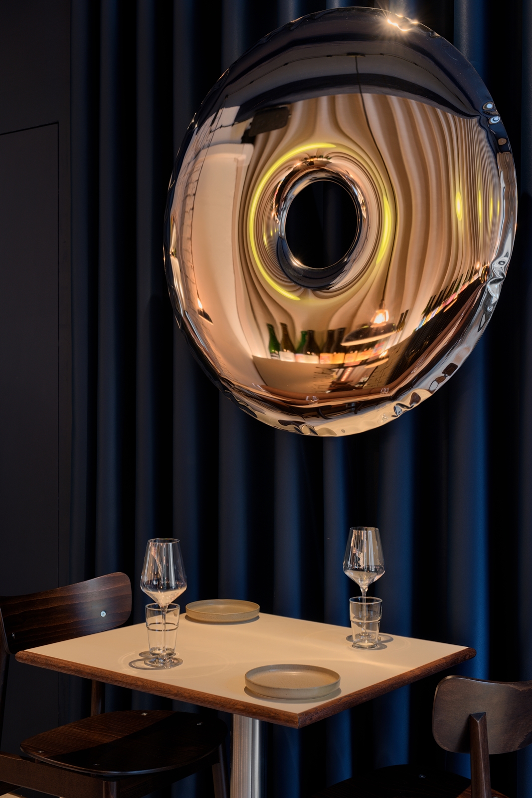 Small dining table with two wooden chairs, set with two plates and two glasses, next to a blue curtain and a large reflective silver donut-shaped balloon.