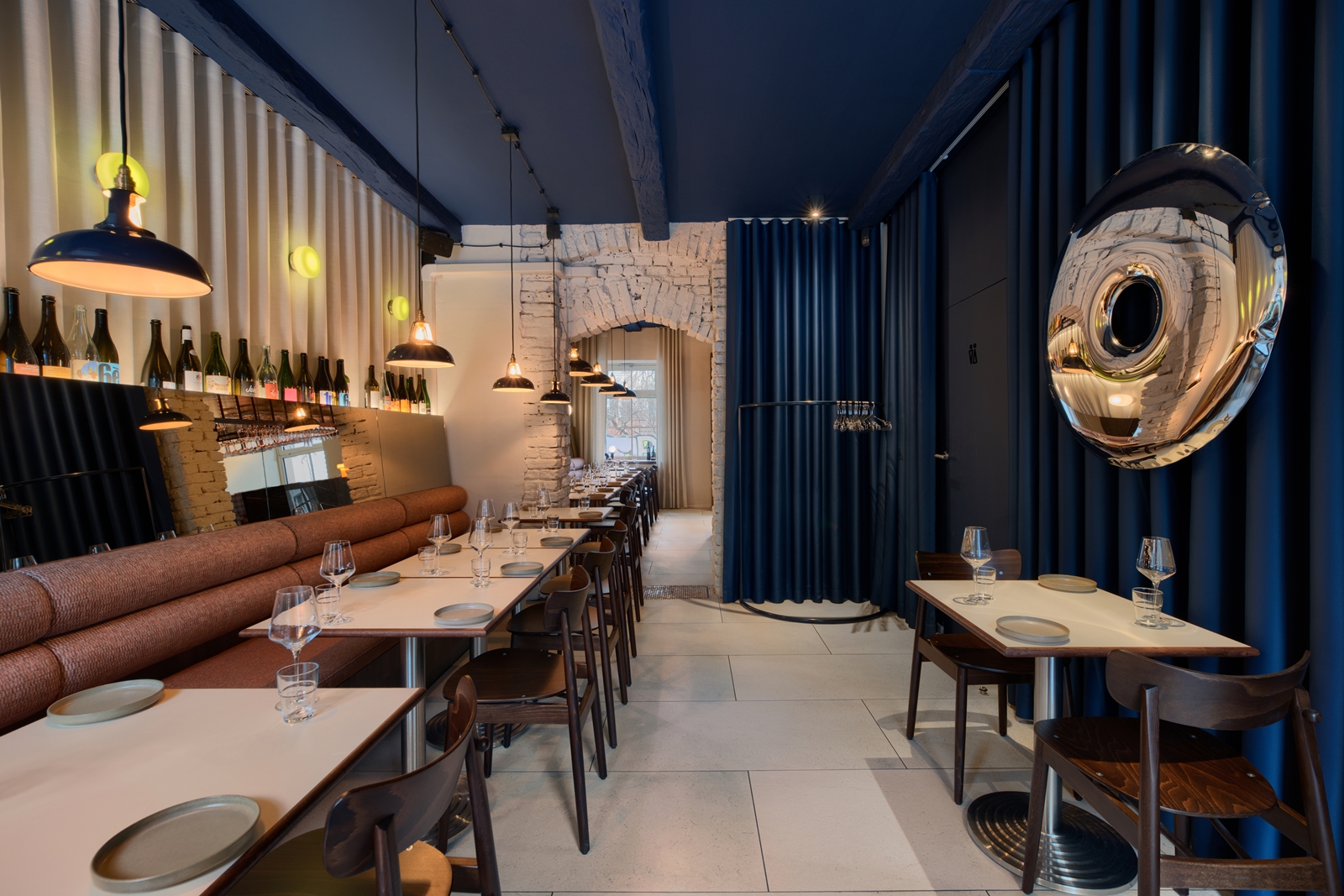 Cozy restaurant interior with a marble bar, wooden chairs, beige cushioned bench seating, and shelves of wine bottles illuminated by pendant lights.