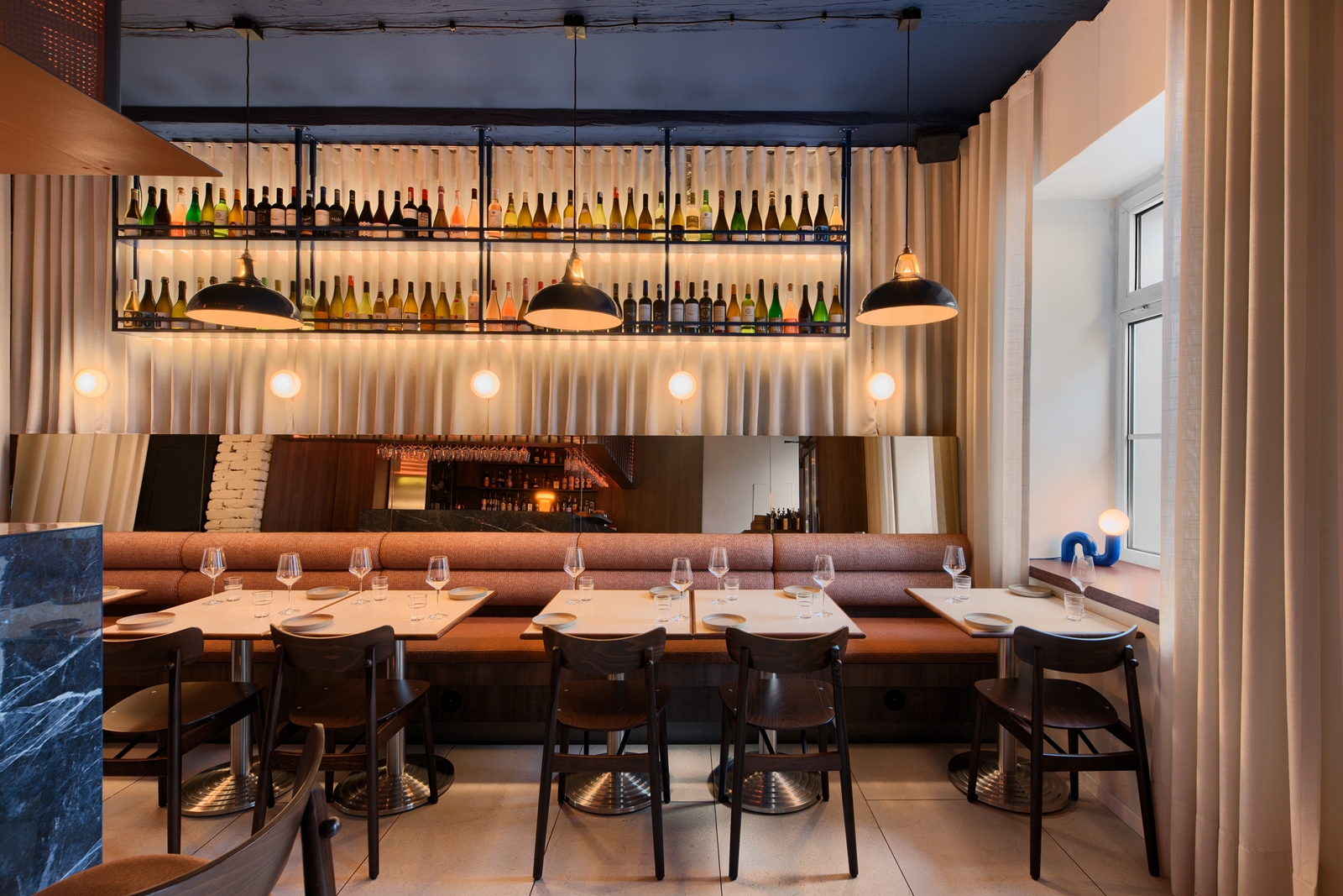 Cozy restaurant interior with a marble bar, wooden chairs, beige cushioned bench seating, and shelves of wine bottles illuminated by pendant lights.
