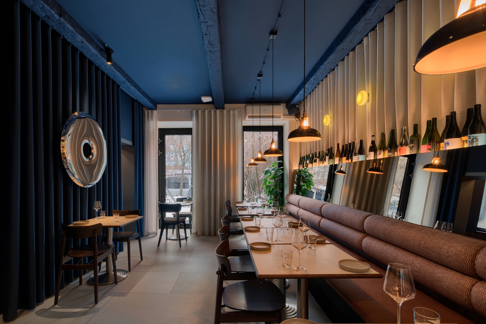 Modern restaurant interior with tables set with plates and glasses, a long cushioned bench, hanging pendant lights, and large windows with curtains.