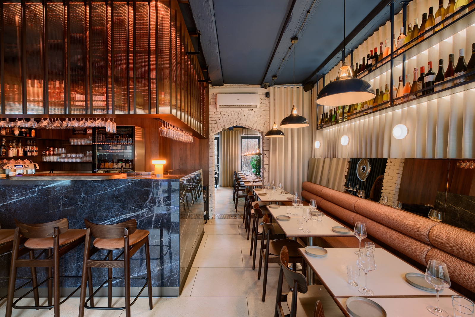 Cozy restaurant interior with cushioned bench seating, wooden chairs, tables set with plates and glasses, and a wine bottle display with hanging lights.