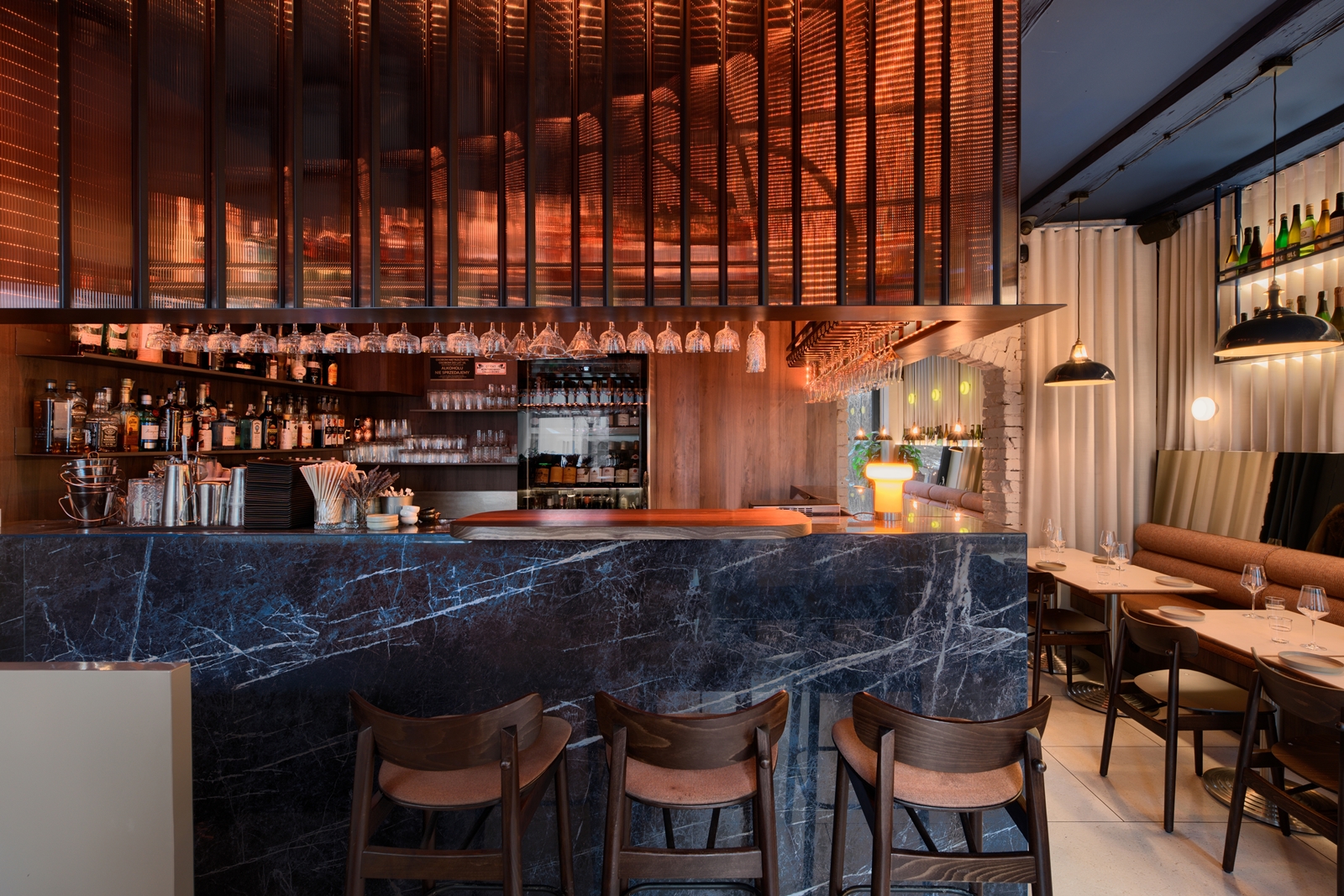 Modern restaurant interior with tables set with plates and glasses, pendant lights, a long cushioned bench, and bottles displayed on a shelf above a mirror.