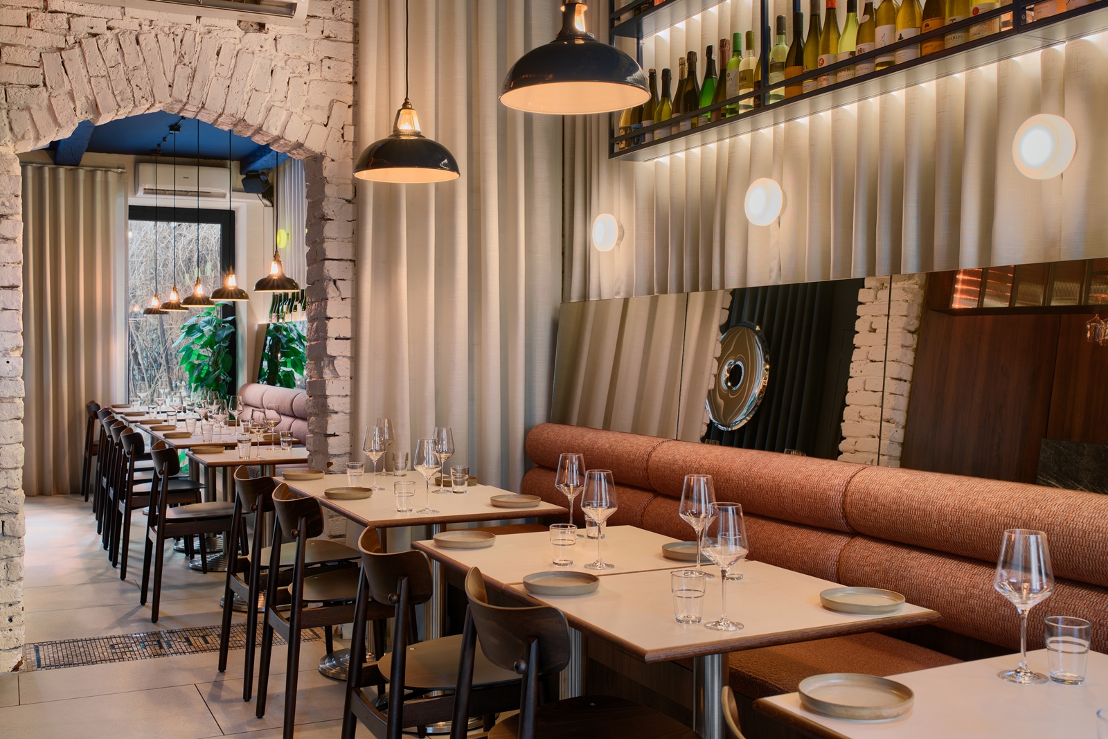 Modern restaurant interior with tables set with plates and glasses, pendant lights, a long cushioned bench, and bottles displayed on a shelf above a mirror.