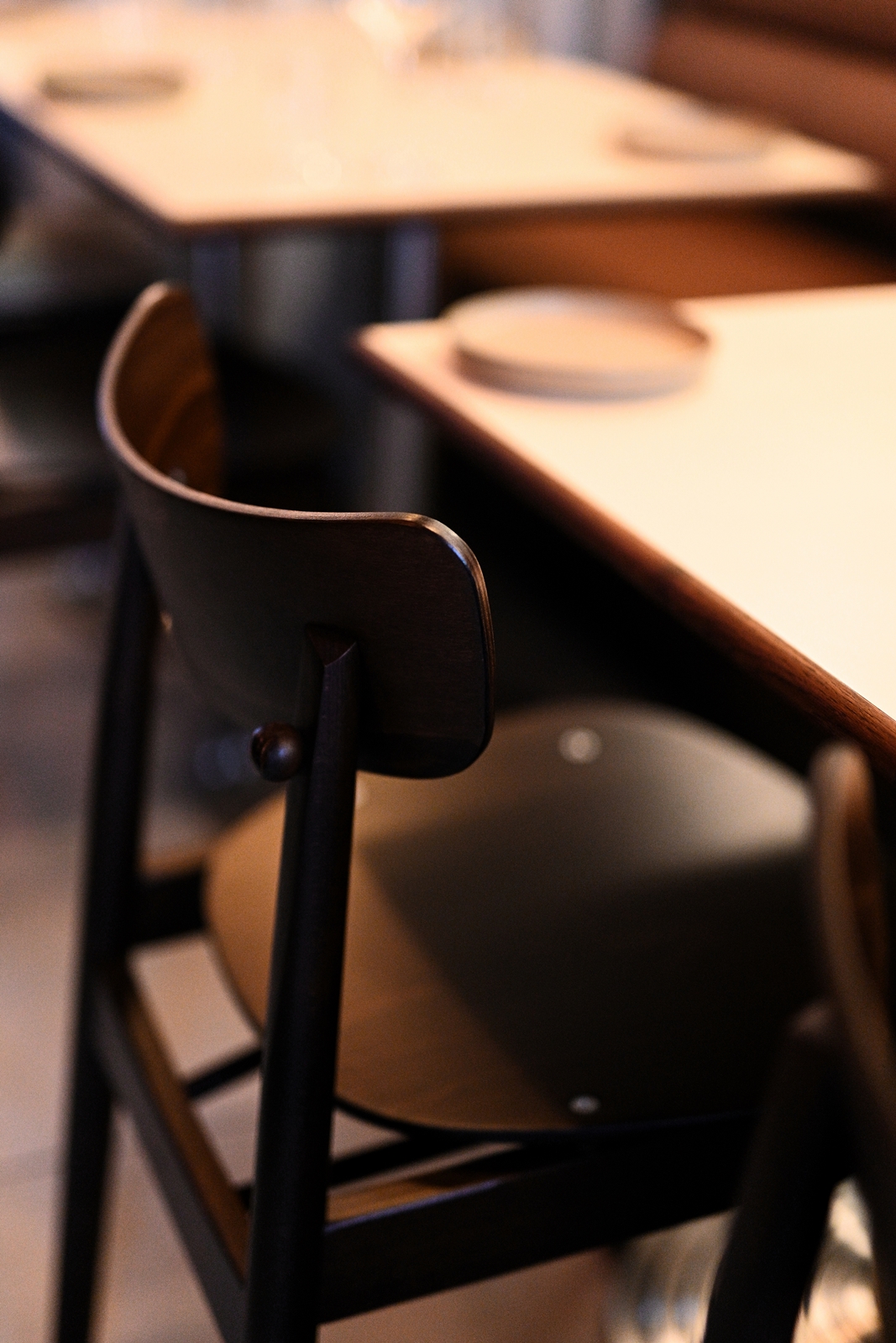 Cozy restaurant corner with two wooden chairs, white tables set with plates and glasses, a cushioned bench, warm lighting, and a window with a modern lamp on the sill.