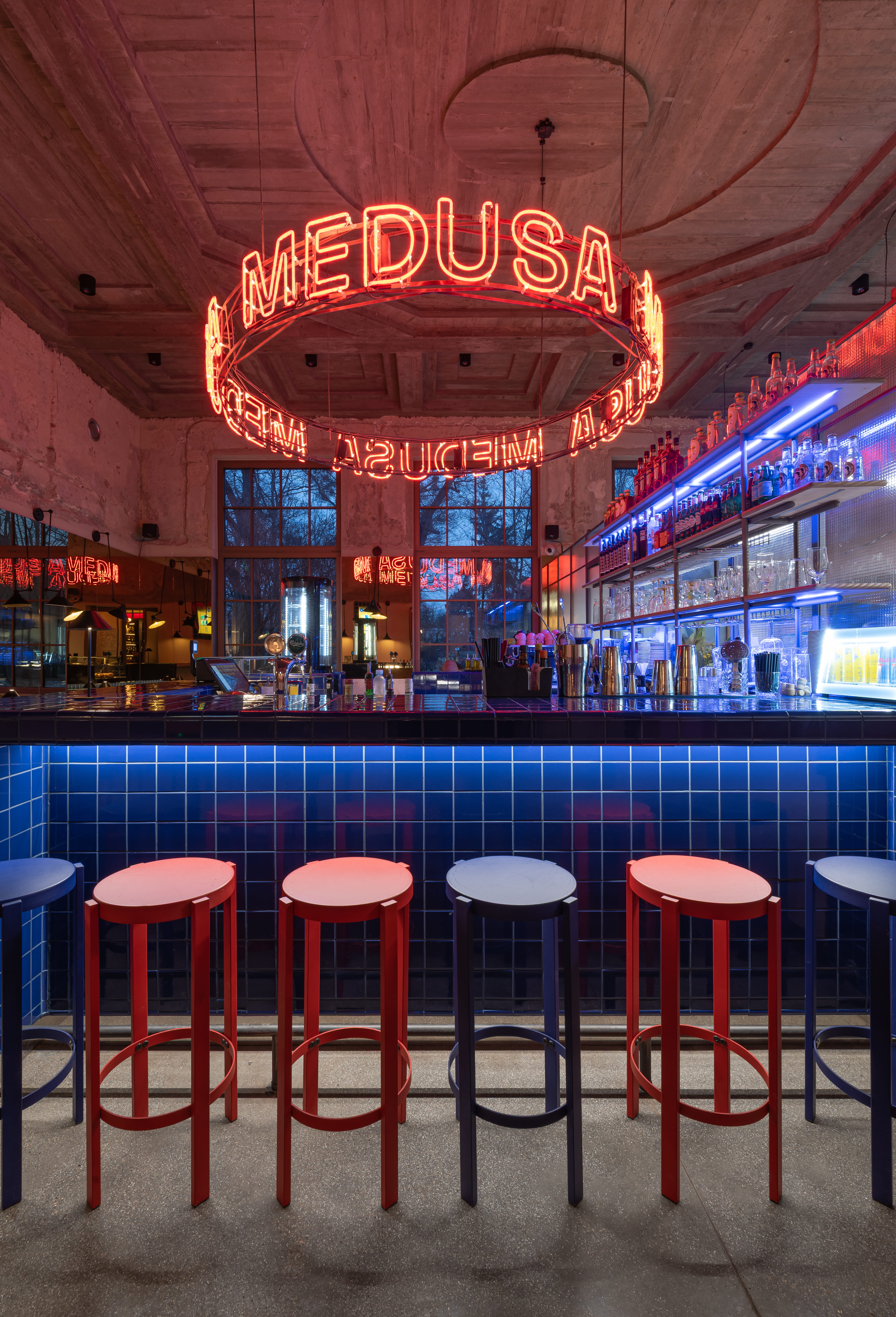 Bar interior with blue tiled counter, red and blue stools, and circular red neon sign reading MEDUSA.