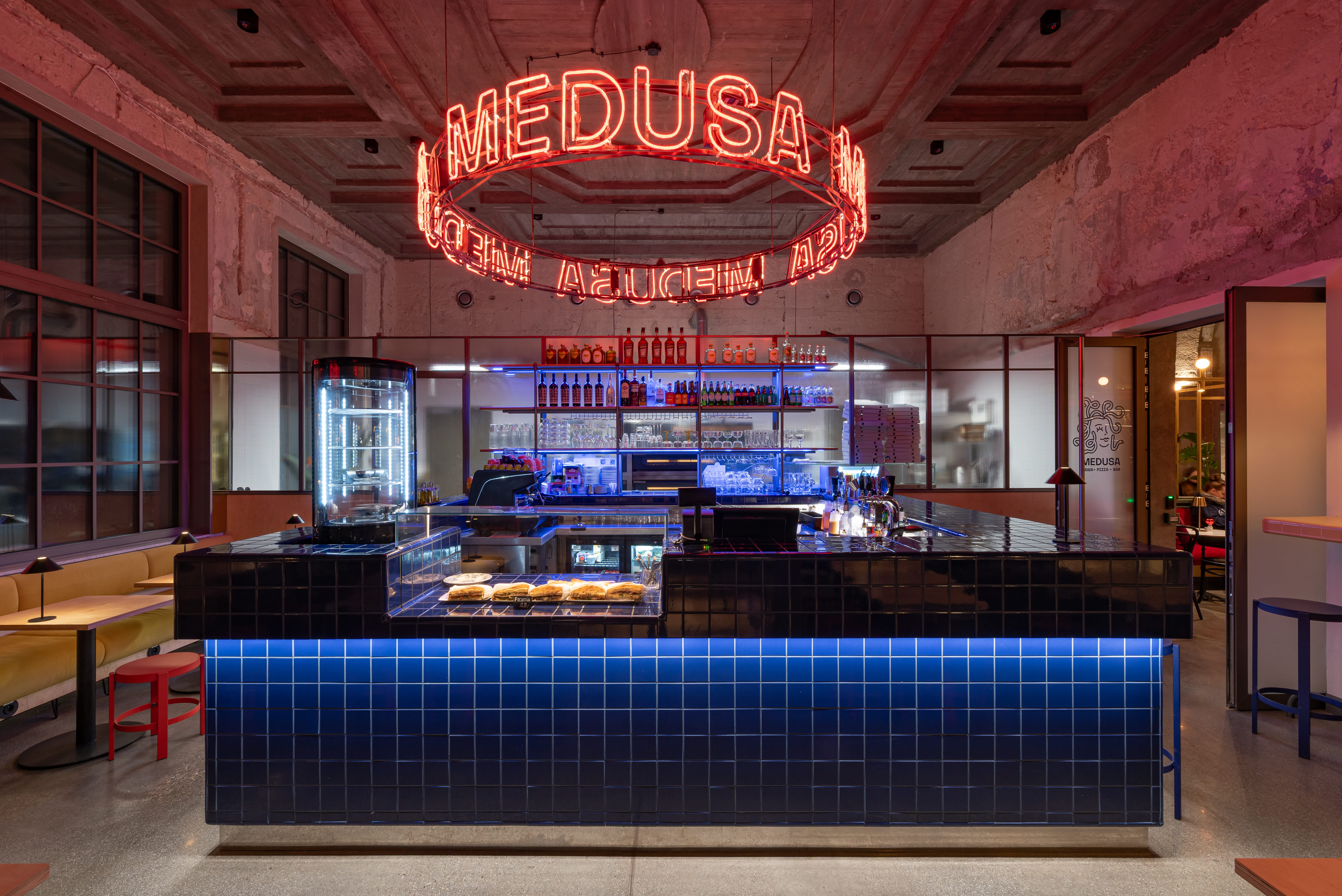 Modern bar with blue tiled counter, neon Medusa sign, and shelves of bottles and glasses.
