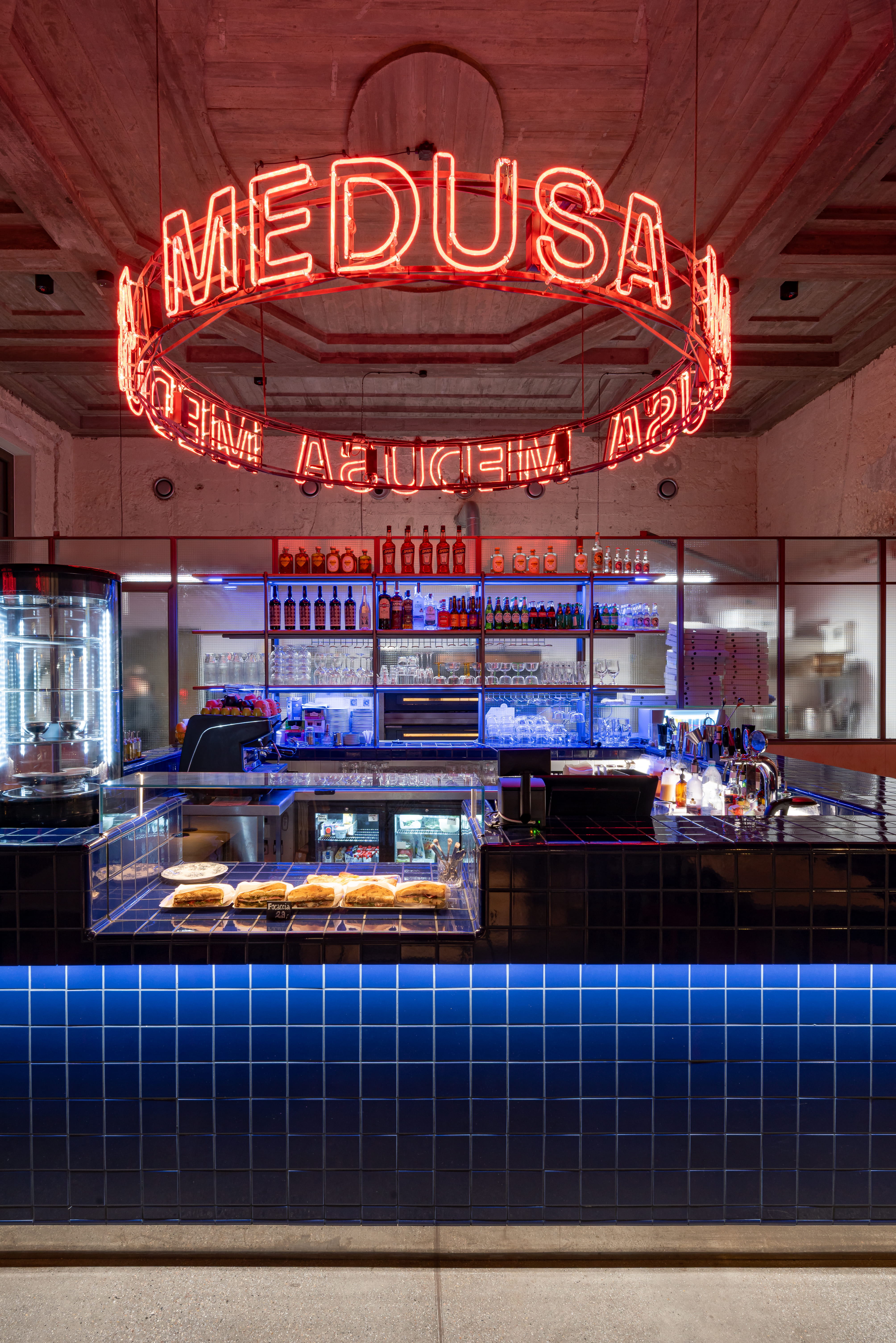 Modern bar counter with blue tile facade, bottles on shelves, and a circular neon sign reading MEDUSA above.