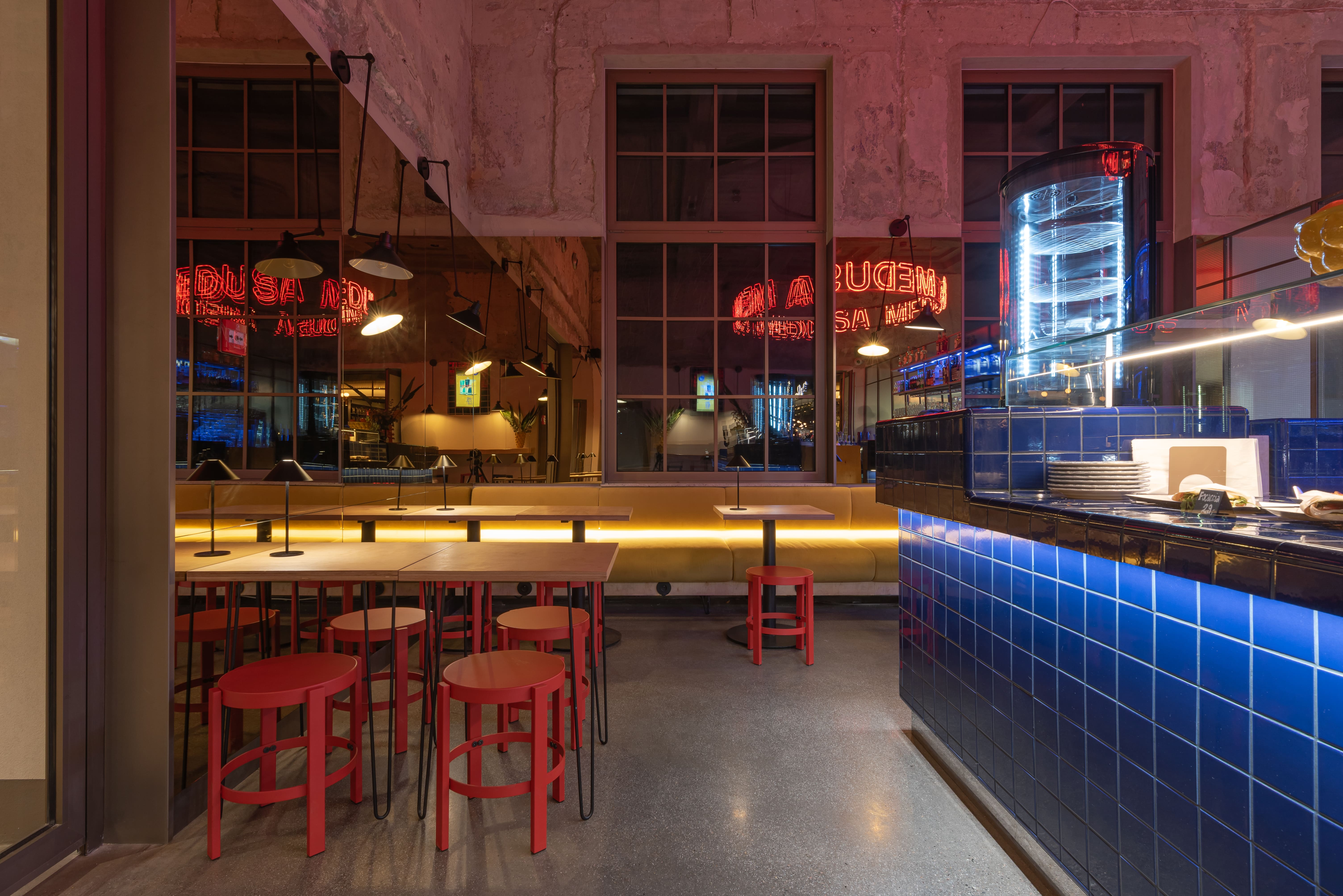 Modern restaurant interior with a central blue-tiled bar, wooden chairs and tables, large windows, and a circular neon light fixture.