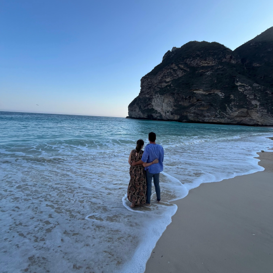 Couple embracing while standing in shallow ocean water on a sandy beach with a large rocky hill in the background under a clear blue sky.