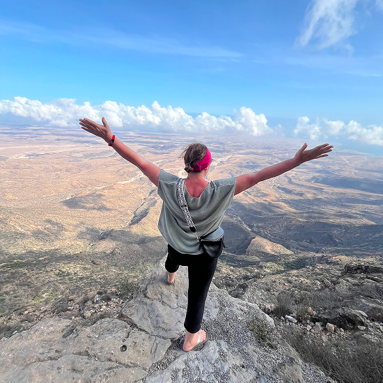 Person standing barefoot on a rocky cliff with arms outstretched overlooking an expansive desert landscape under a blue sky with clouds.