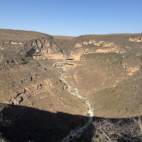 Arid canyon landscape with rocky cliffs, a dry riverbed, and clear blue sky.