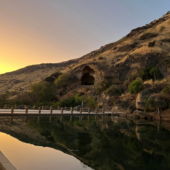 Sunset over a hillside with a large cave entrance above a wooden walkway and calm reflective water.