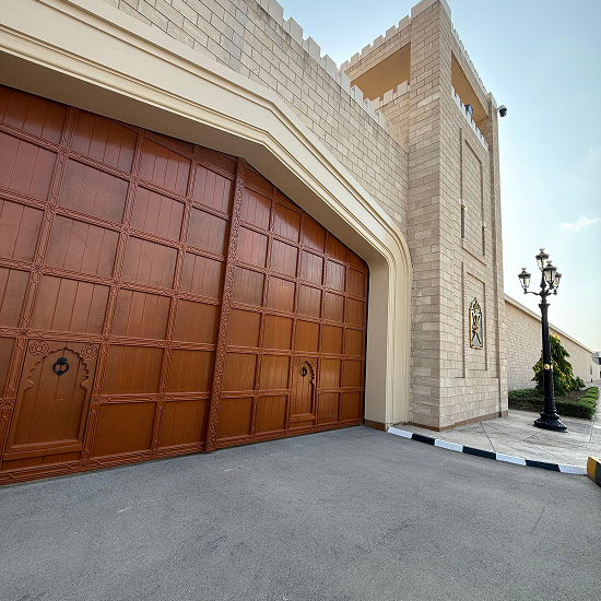 Large light brown stone gate tower with a tall wooden gate and a decorative metal wall fixture, beside a black street lamp and paved road.