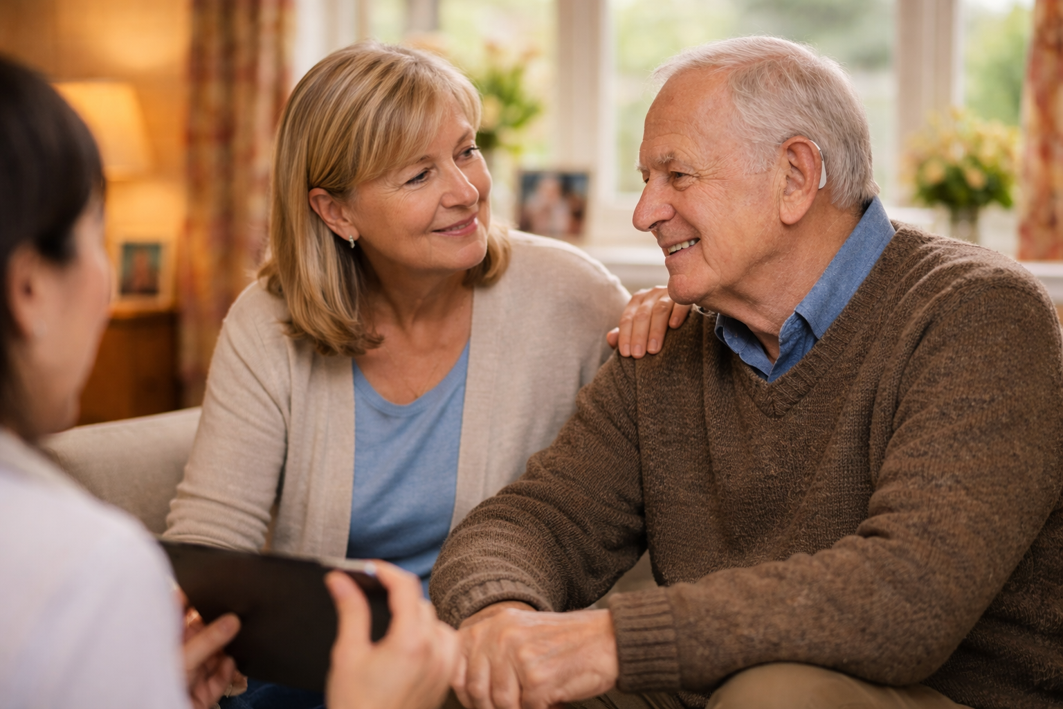 Elderly father smiling during a home hearing consultation, with his daughter seated beside him in a warmly lit living room.