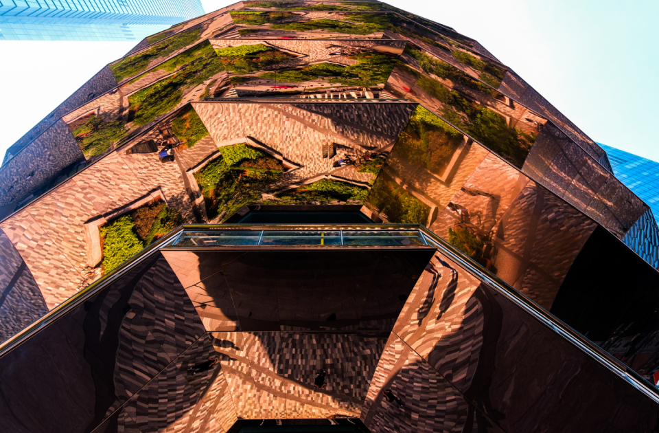 Upward view of the Vessel, a mirrored, honeycomb-structured architectural landmark reflecting its staircases and surrounding greenery.
