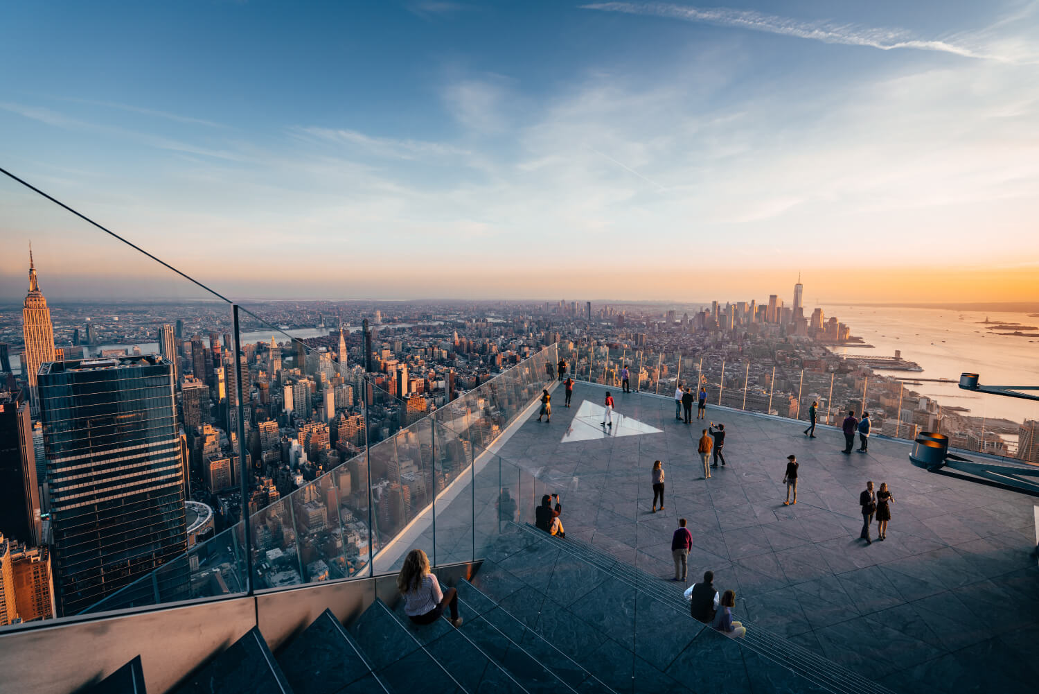 View of New York City skyline at sunset from a glass-paneled observation deck with people walking and sitting.