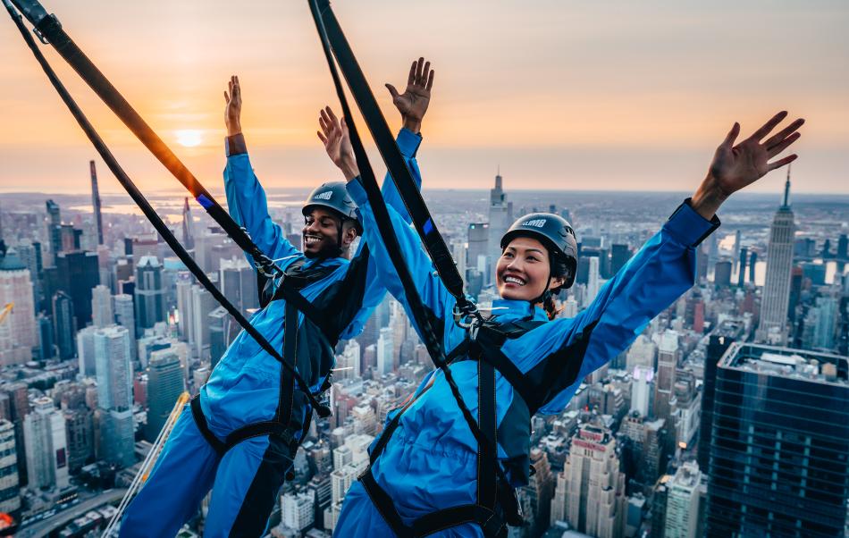 Two people in blue harnesses and helmets smiling with arms raised while suspended above a city skyline at sunset.