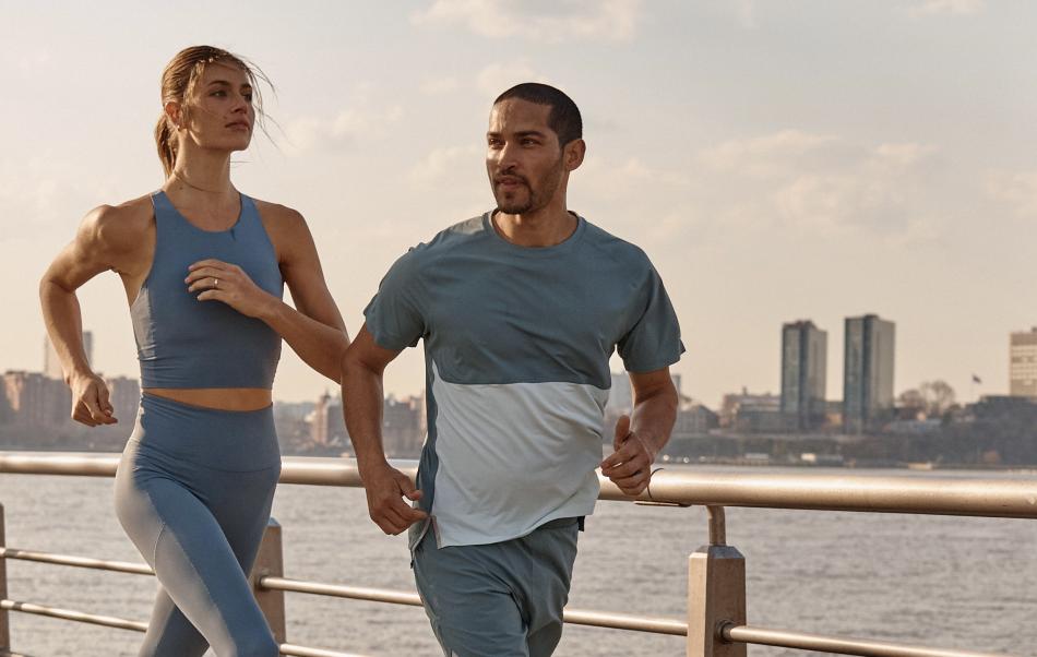 A man and a woman jogging outdoors along a waterfront with city buildings in the background.