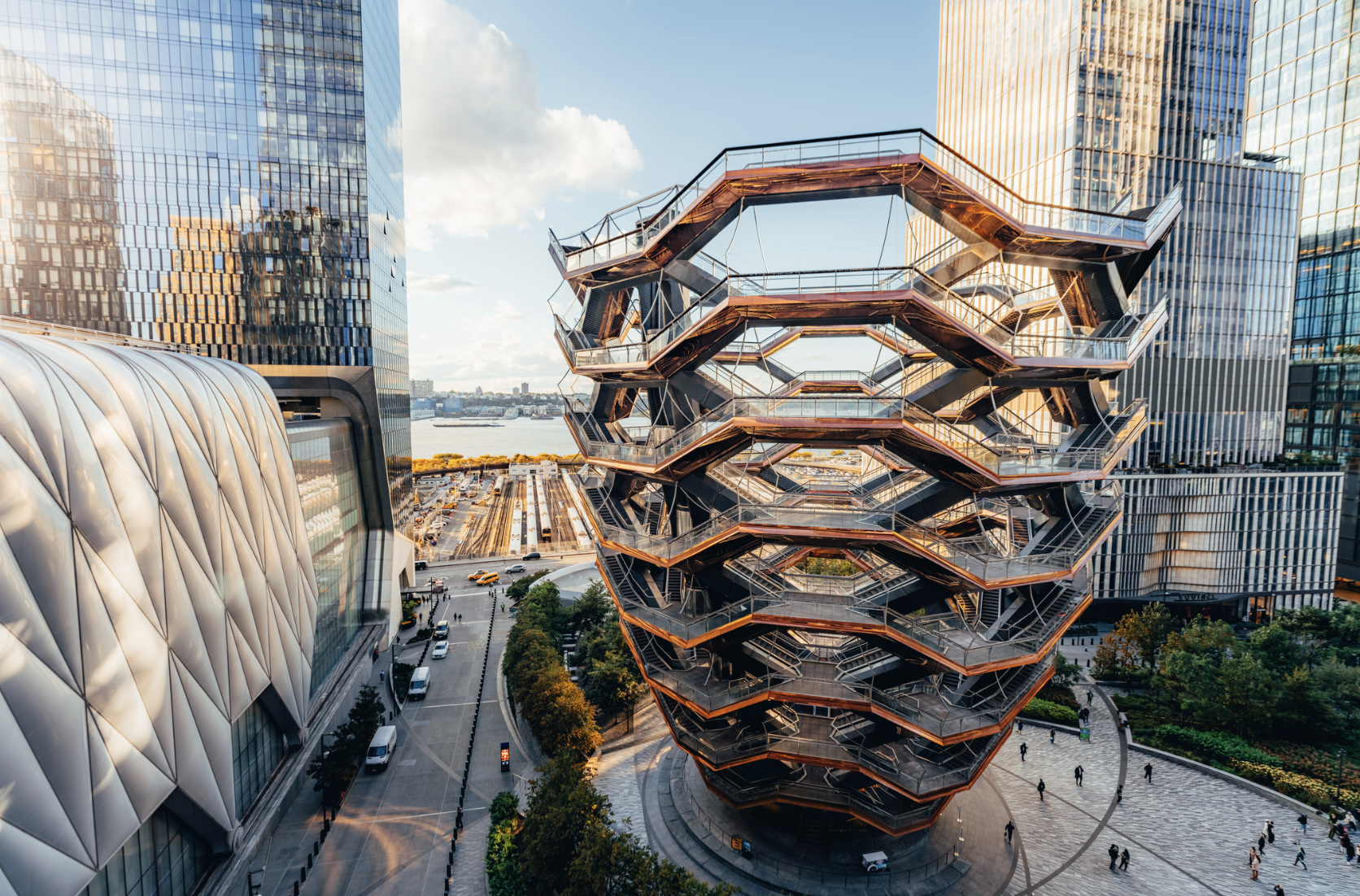 The Vessel, a honeycomb-like structure with interconnected staircases in an urban plaza surrounded by modern skyscrapers and greenery at sunset.