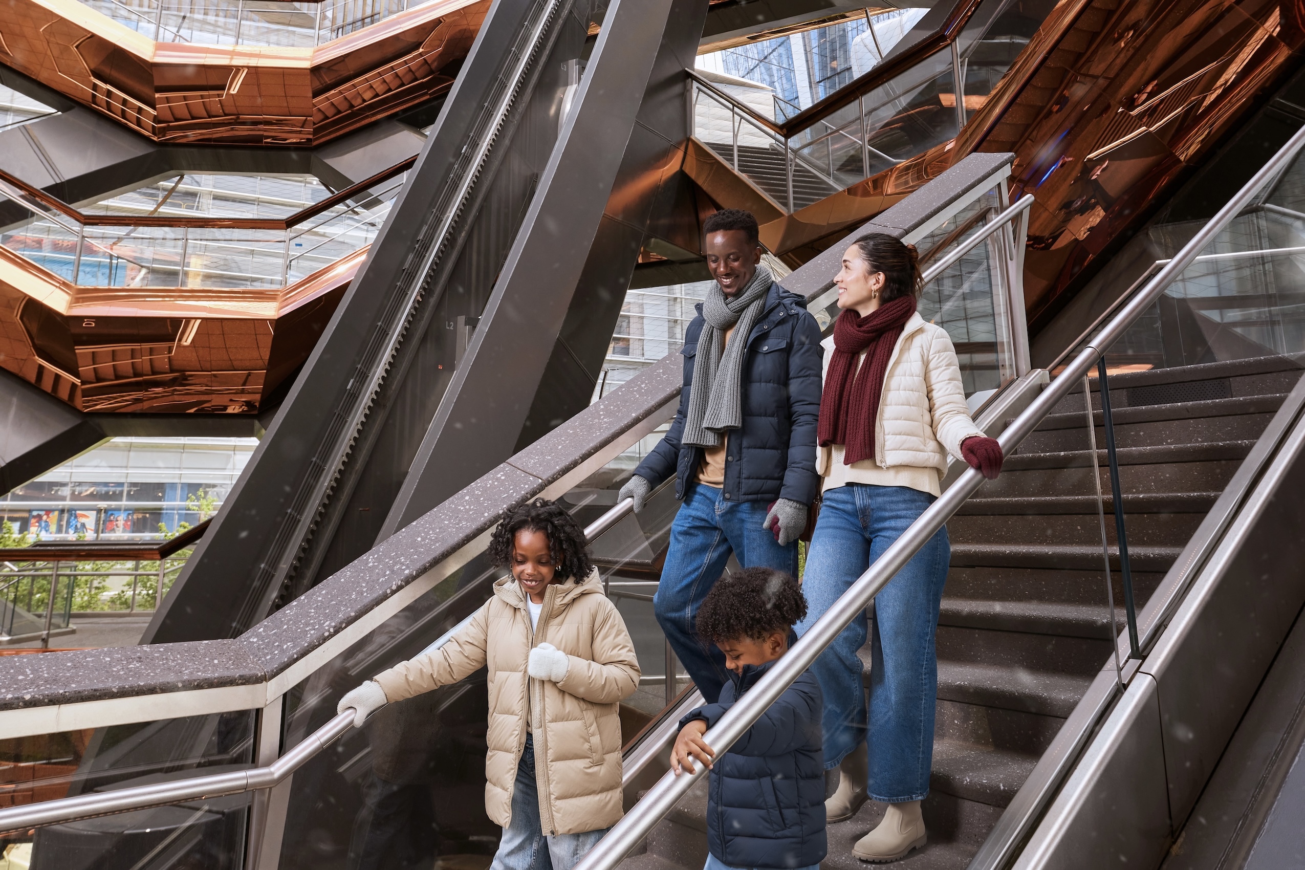 Family of four dressed in winter clothes walking down stairs inside a modern building with copper geometric design.