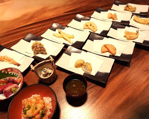A Japanese omakase meal with various tempura dishes on rectangular plates, a bowl of rice topped with tempura, a sashimi bowl, a small cup with a side dish, and a bowl of green soup on a wooden table.