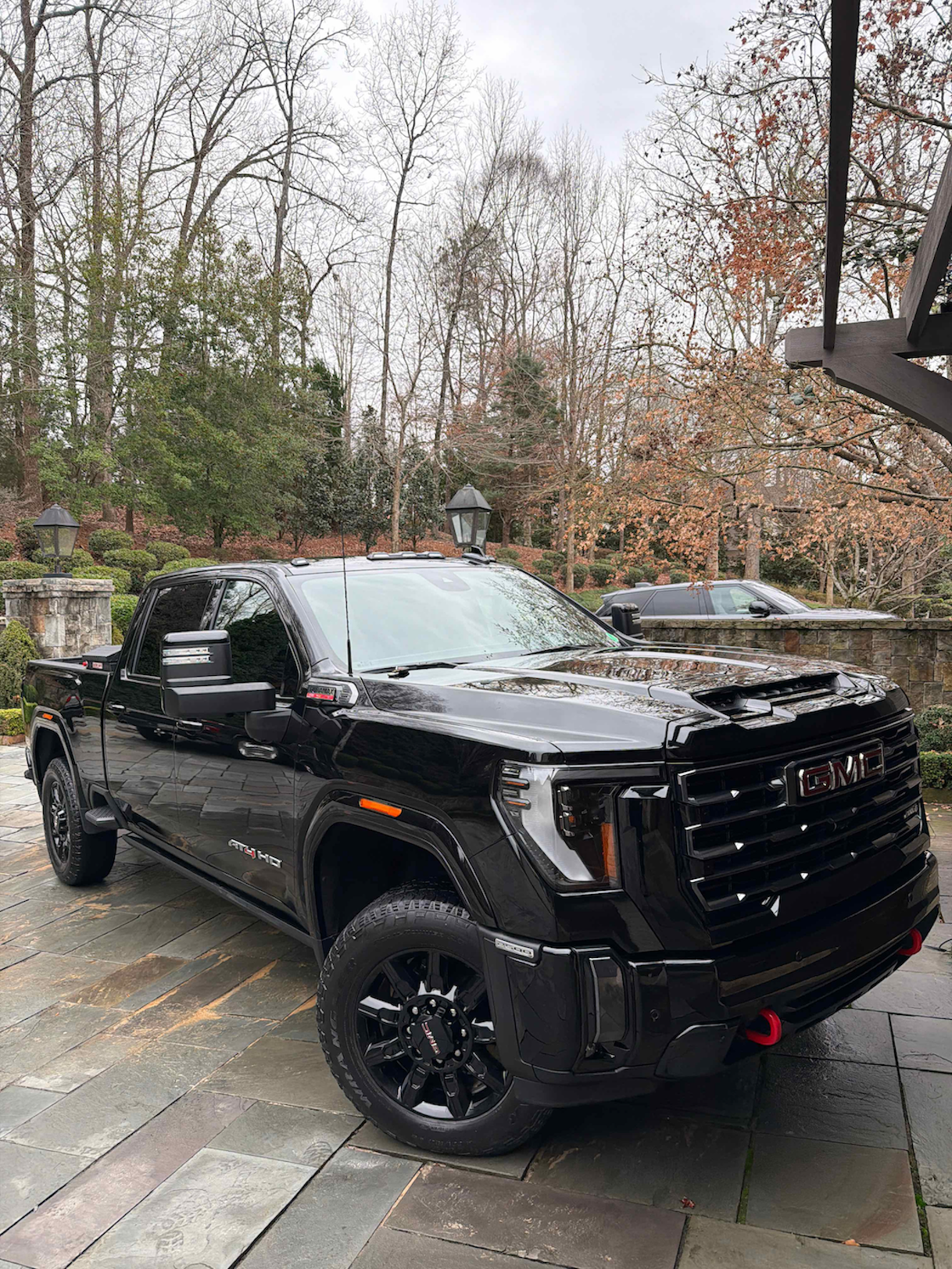 Black GMC AT4 HD pickup truck parked on a stone driveway surrounded by leafless trees and landscaping.