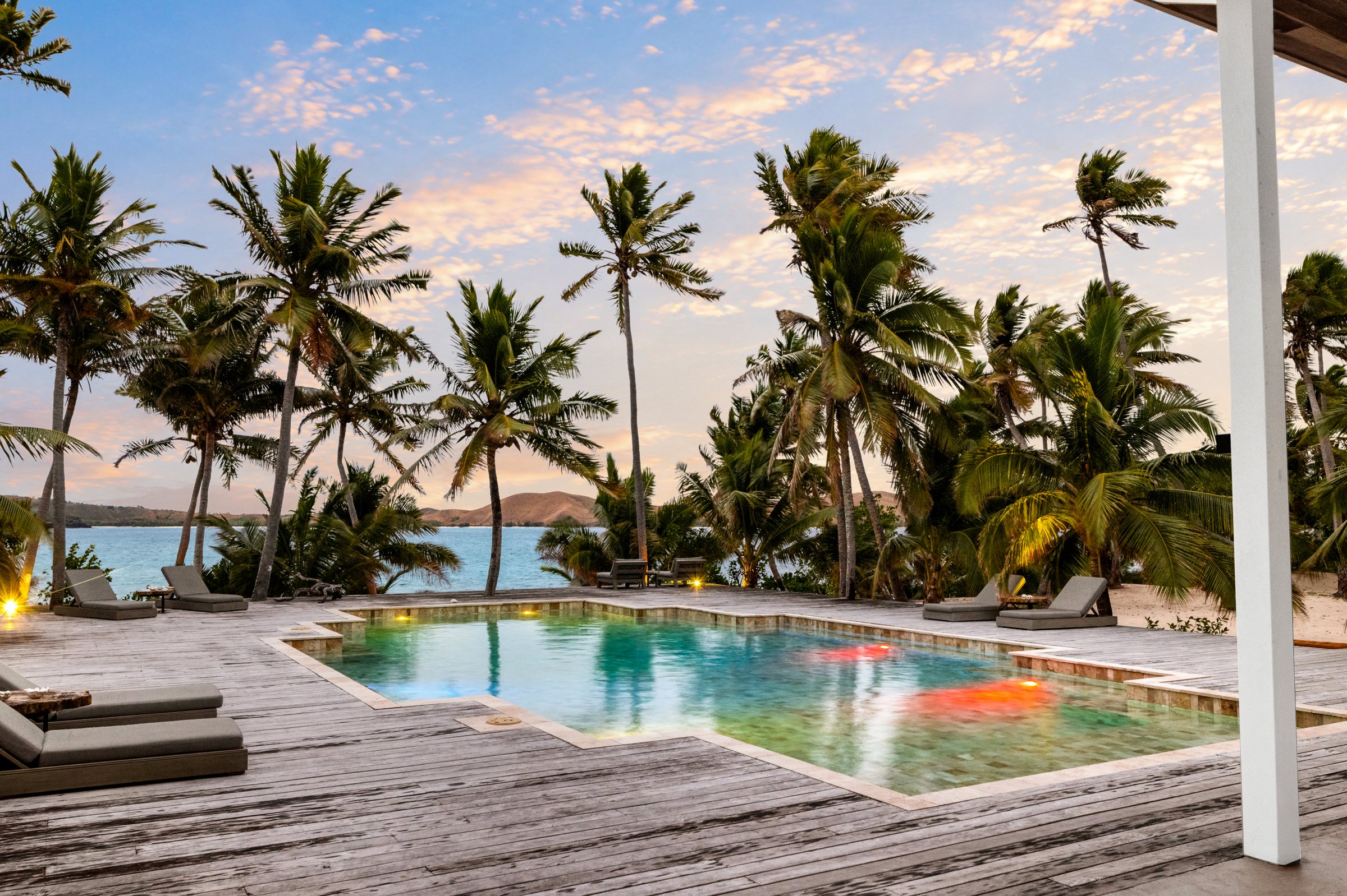 Pool glowing at dusk beneath palms, with loungers along the deck and hills across the water