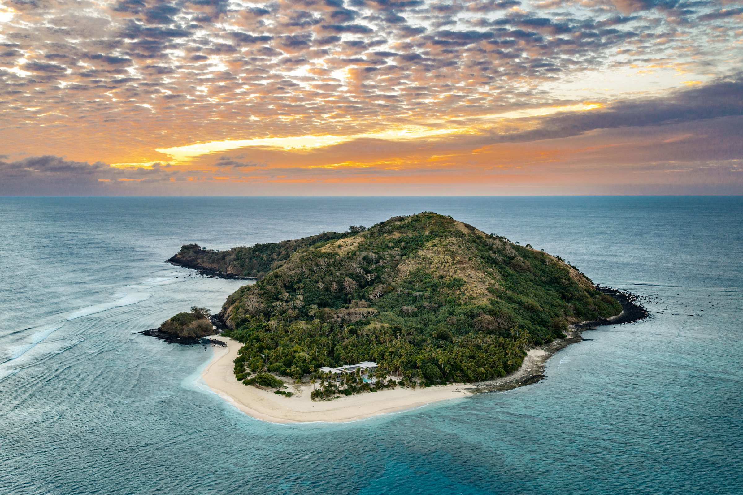 Aerial view of the private island at sunset, with the residence set above a crescent of white sand