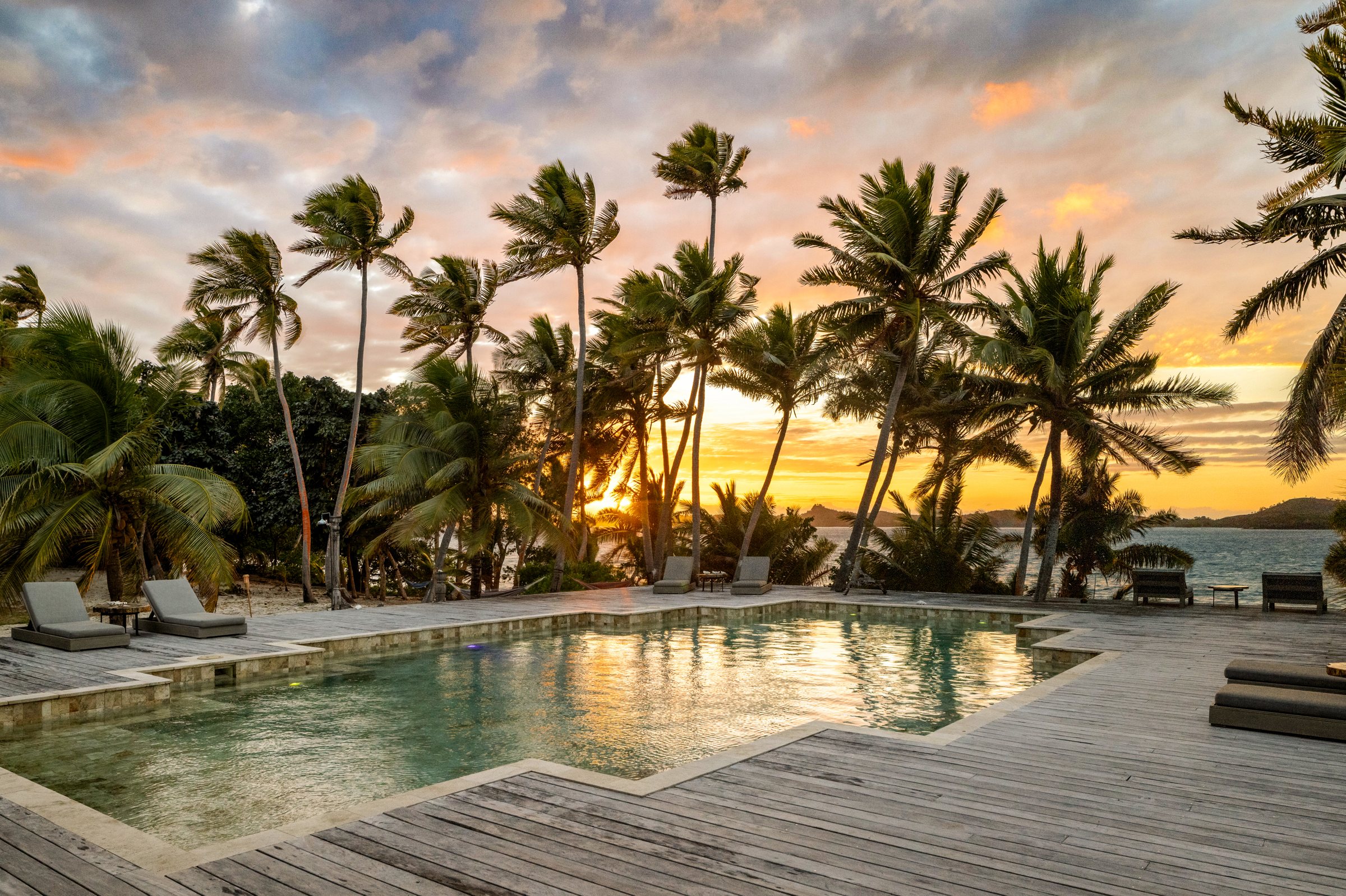 Pool and timber deck at sunset, framed by tall coconut palms and the bay