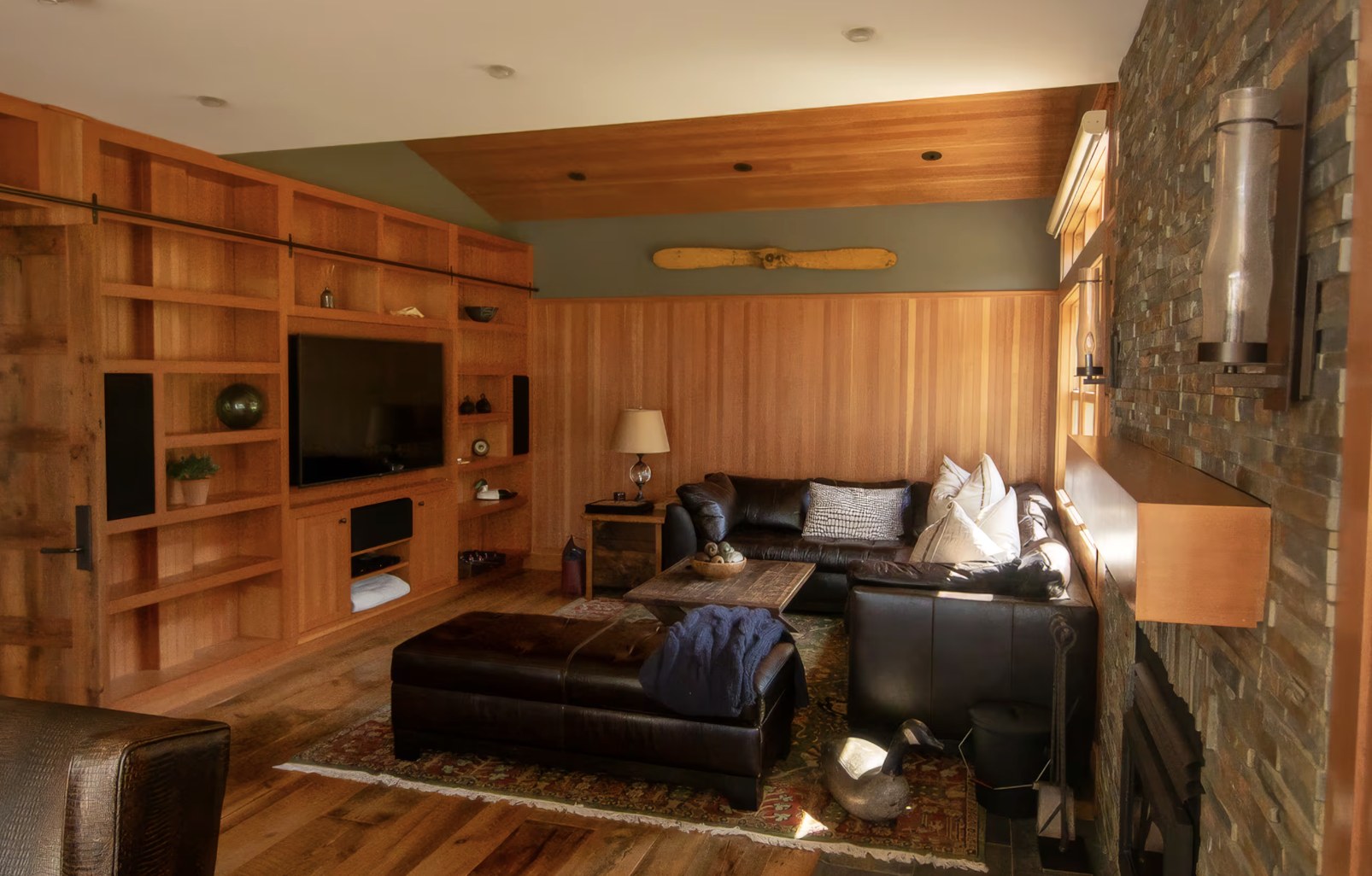 Den with Douglas fir paneling, built-in media shelves, a leather sectional, and a stone fireplace