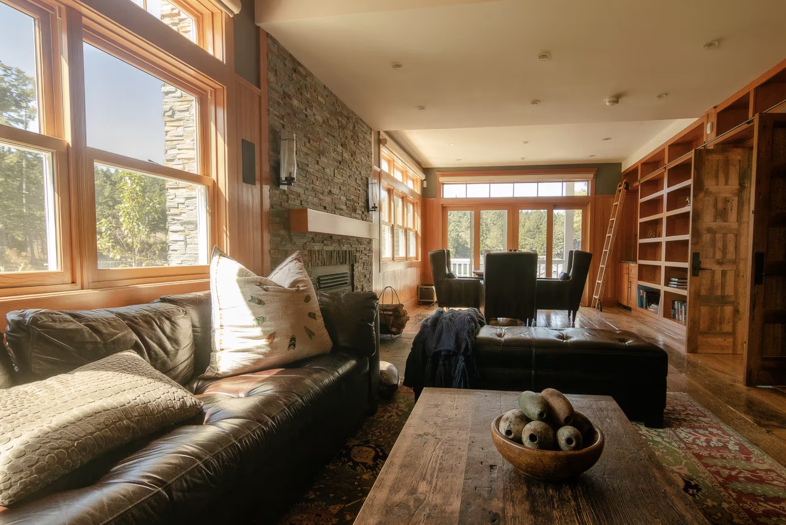 Living room with a leather sofa, stacked stone fireplace, and tall windows onto the trees