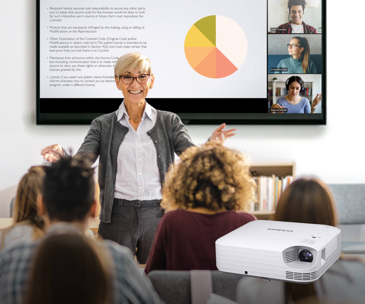 Smiling woman giving a presentation to an audience with a pie chart and text on screen, and three people joining virtually in video call, with a Casio projector in foreground.