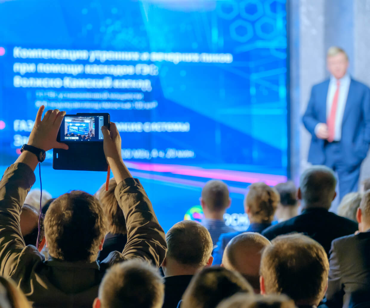 Audience watching a speaker in a blue suit and red tie at a conference while one person records the presentation on a tablet.