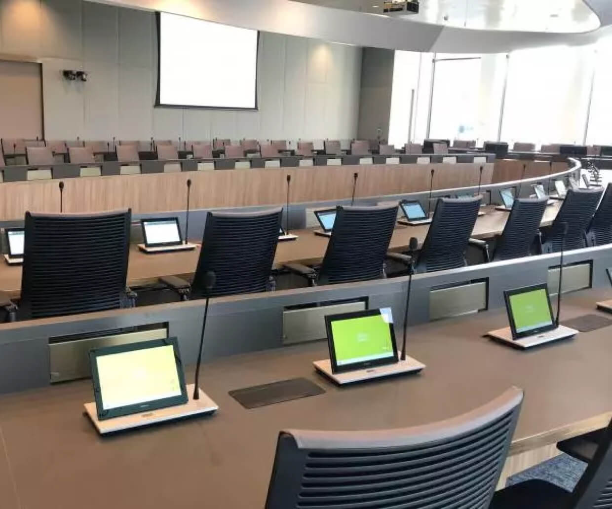 Modern conference room with curved tables, black office chairs, built-in microphones, and tablet screens at each seat.
