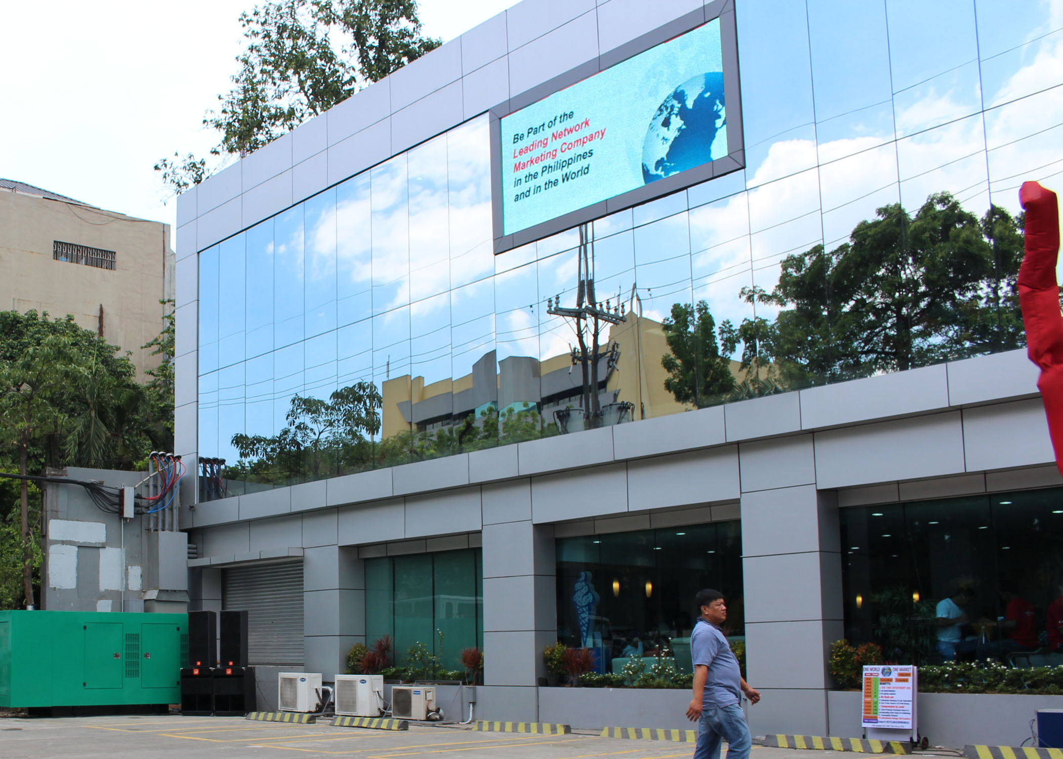 Modern building with glass panels reflecting sky and trees, a man walking in front of it.