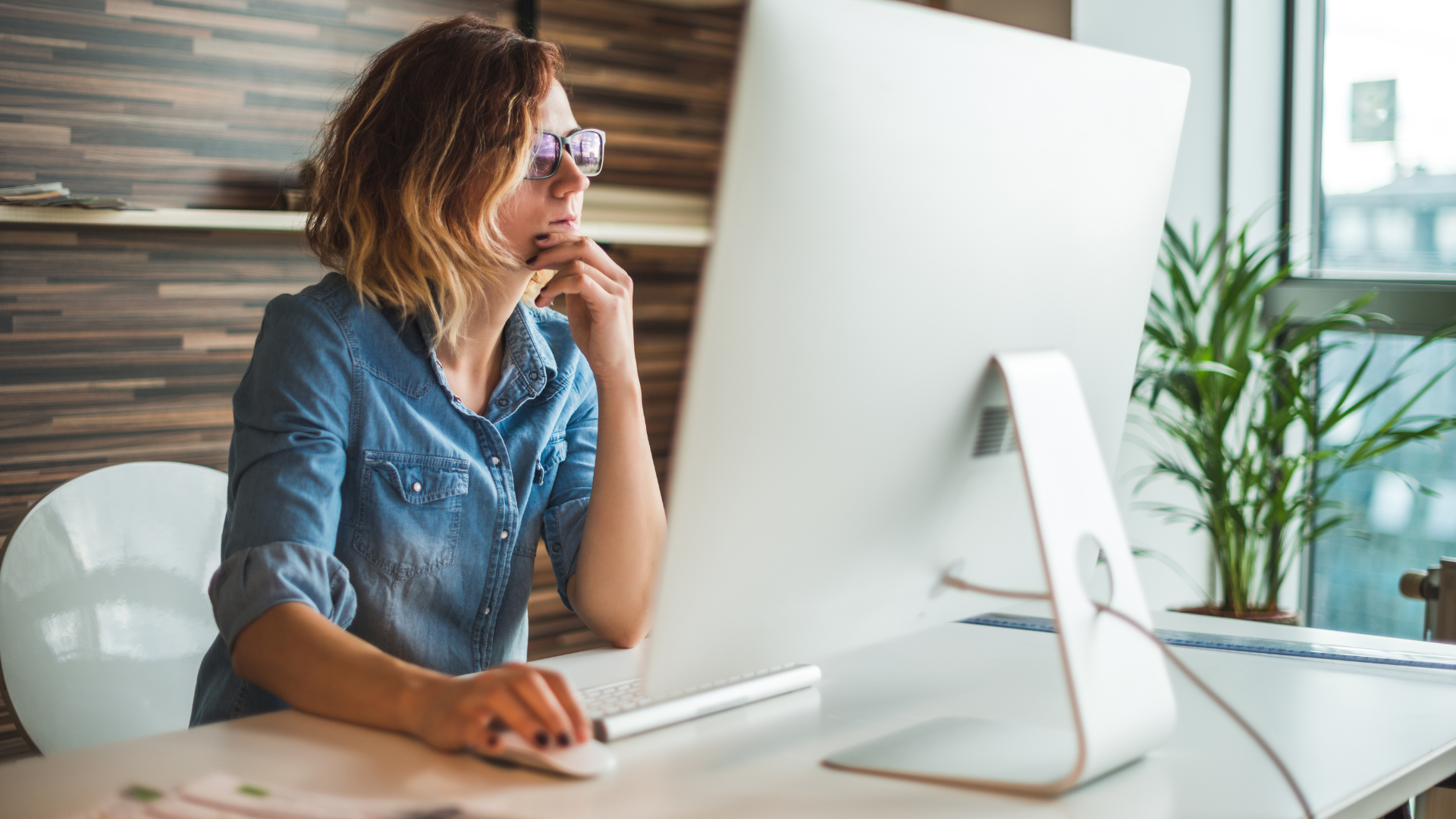 A young woman with shoulder-length brown hair and glasses sitting at a white iMac desktop in a modern office with wood-plank wall and houseplant, thoughtfully reviewing a landscape company website on screen