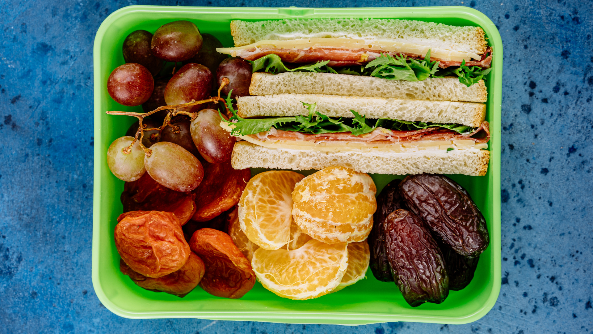 Top-down view of a green plastic lunch box packed with a triple-decker sandwich on white bread, red grapes on stems, dried apricots, peeled mandarin orange segments, and medjool dates on a dark blue speckled surface