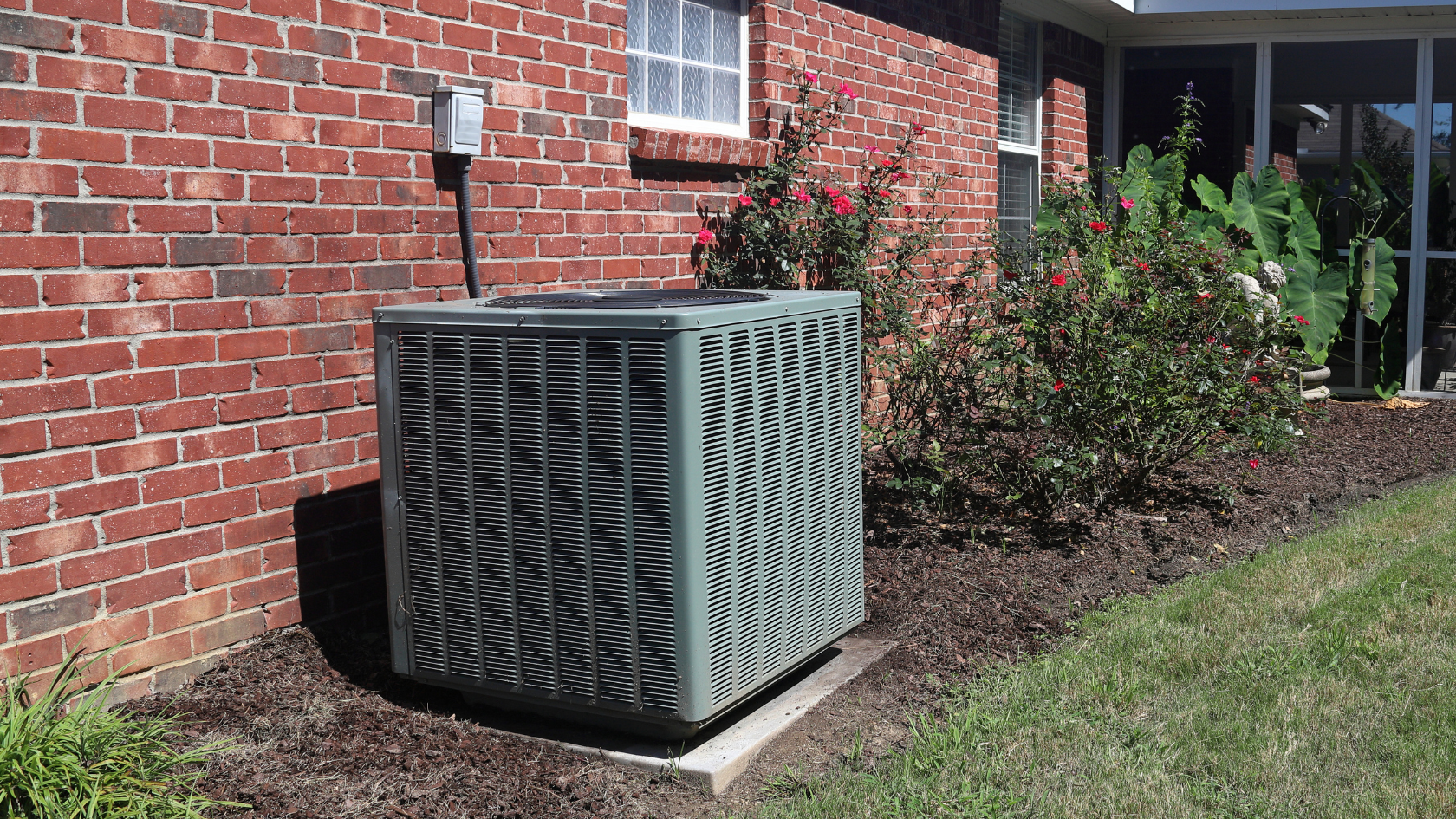 Residential outdoor HVAC condenser on a concrete pad against a red brick wall with an electrical disconnect box and a flowering rose bush, illustrating the kind of equipment install that anchors HVAC pricing conversations for homeowners and contractors.
