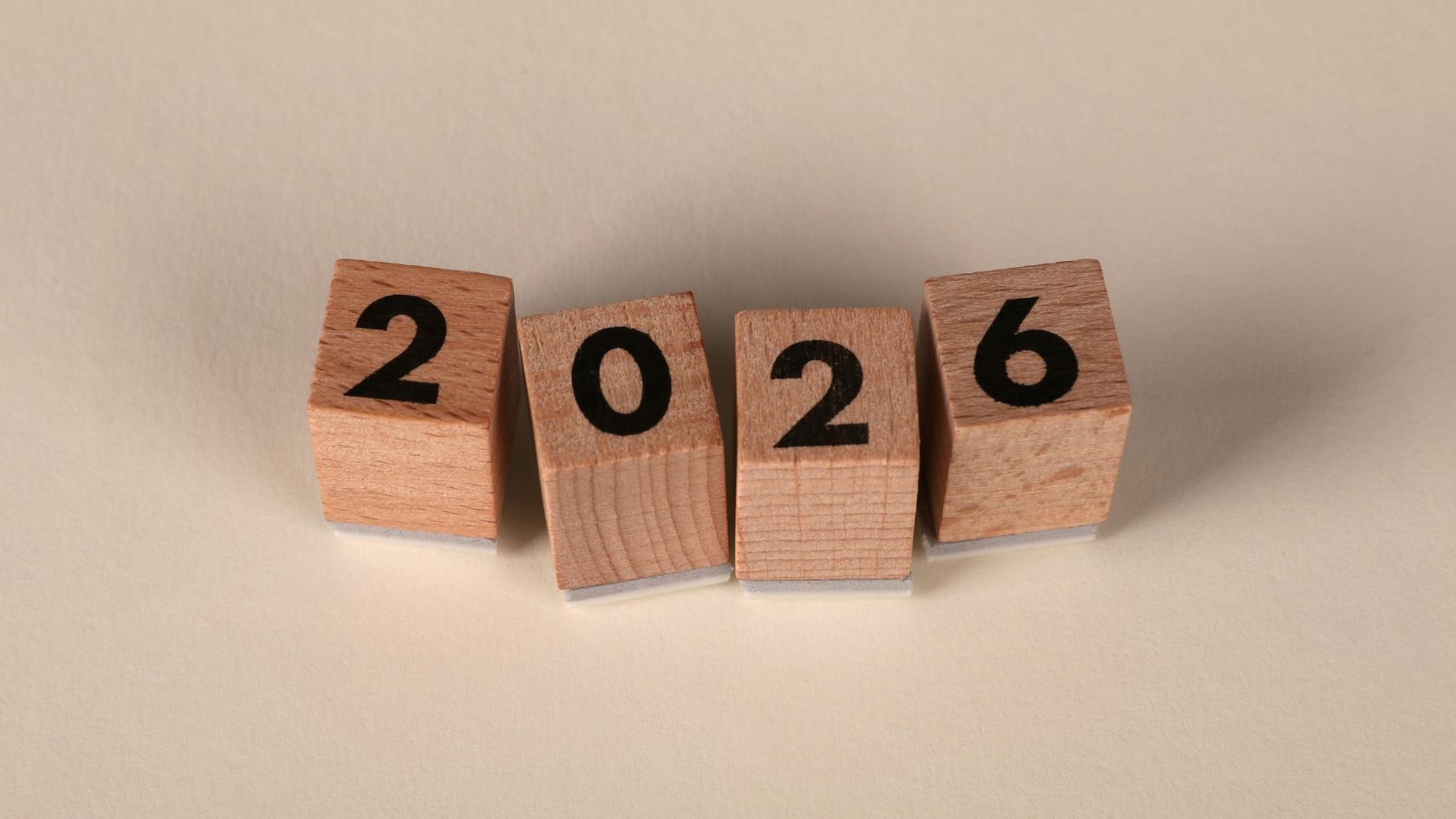 Four small wooden blocks arranged in a row on a warm beige paper background, each block showing one black-printed digit that together spell the year 2026, with the middle two blocks tilted slightly forward to display the numerals clearly.