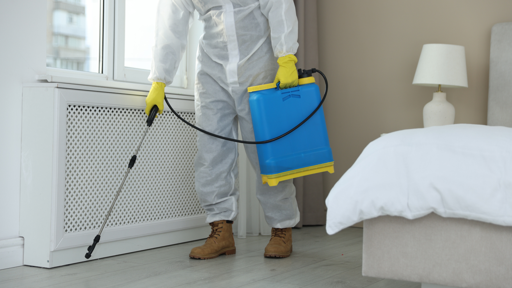 Pest control technician in white Tyvek coveralls and yellow gloves operating a blue and yellow backpack sprayer near a window radiator inside a residential bedroom, the indoor winter treatment that keeps pest control crews booked through the offseason.