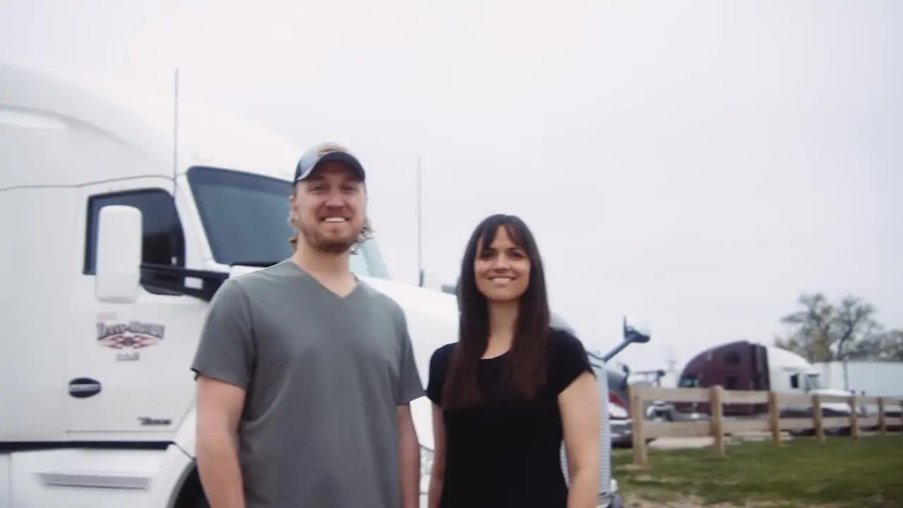 Smiling man and woman standing in front of parked semi trucks outdoors.