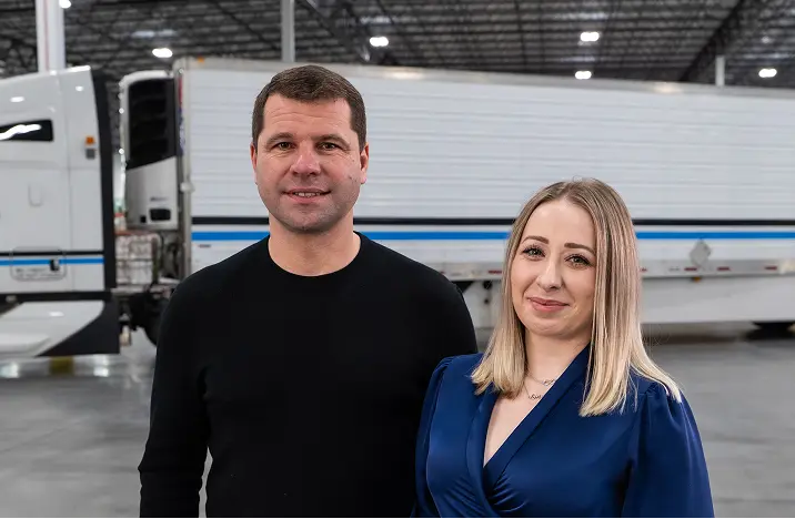 Man and woman standing inside a warehouse in front of a white truck trailer.