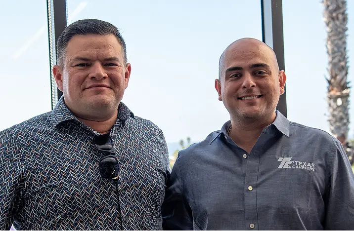 Two men smiling indoors near windows, one wearing a patterned shirt with sunglasses hanging and the other wearing a Texas Carriers shirt.