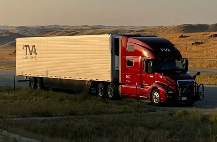Red TVA Logistics Inc. semi-truck parked on the roadside in a rural area with rolling hills in the background at sunset.