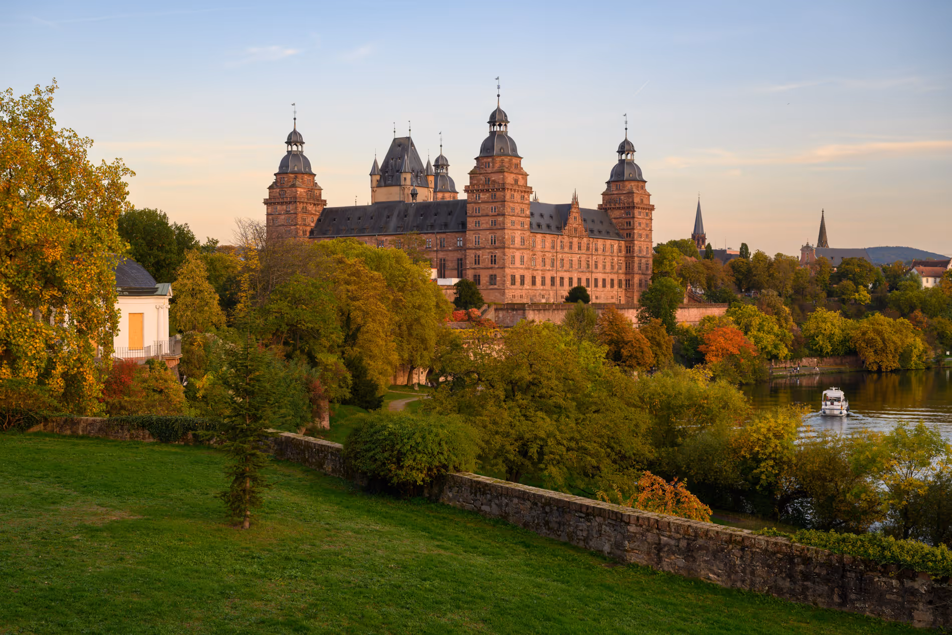 Schloss Johannisburg Aschaffenburg bei Sonnenuntergang mit umliegenden Bäumen und einem Boot auf dem Fluss.