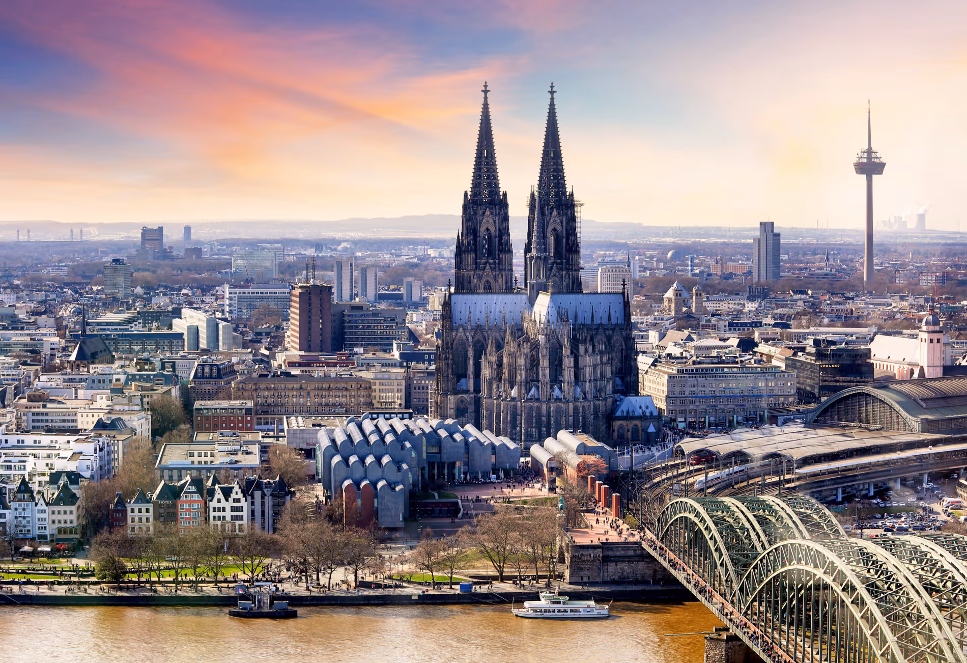 Kölner Dom mit Hohenzollernbrücke und Stadtpanorama bei Sonnenuntergang.