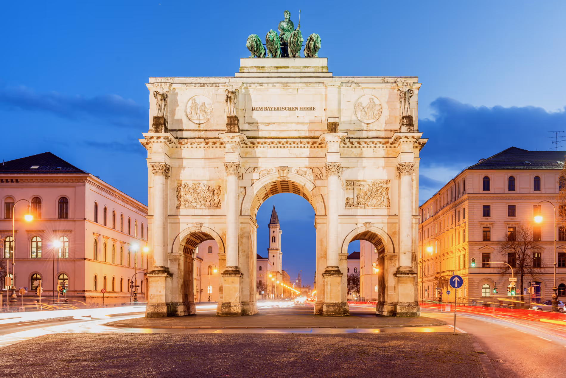 Die beleuchtete Siegestor in München bei Abenddämmerung mit Verkehr und historischen Gebäuden.