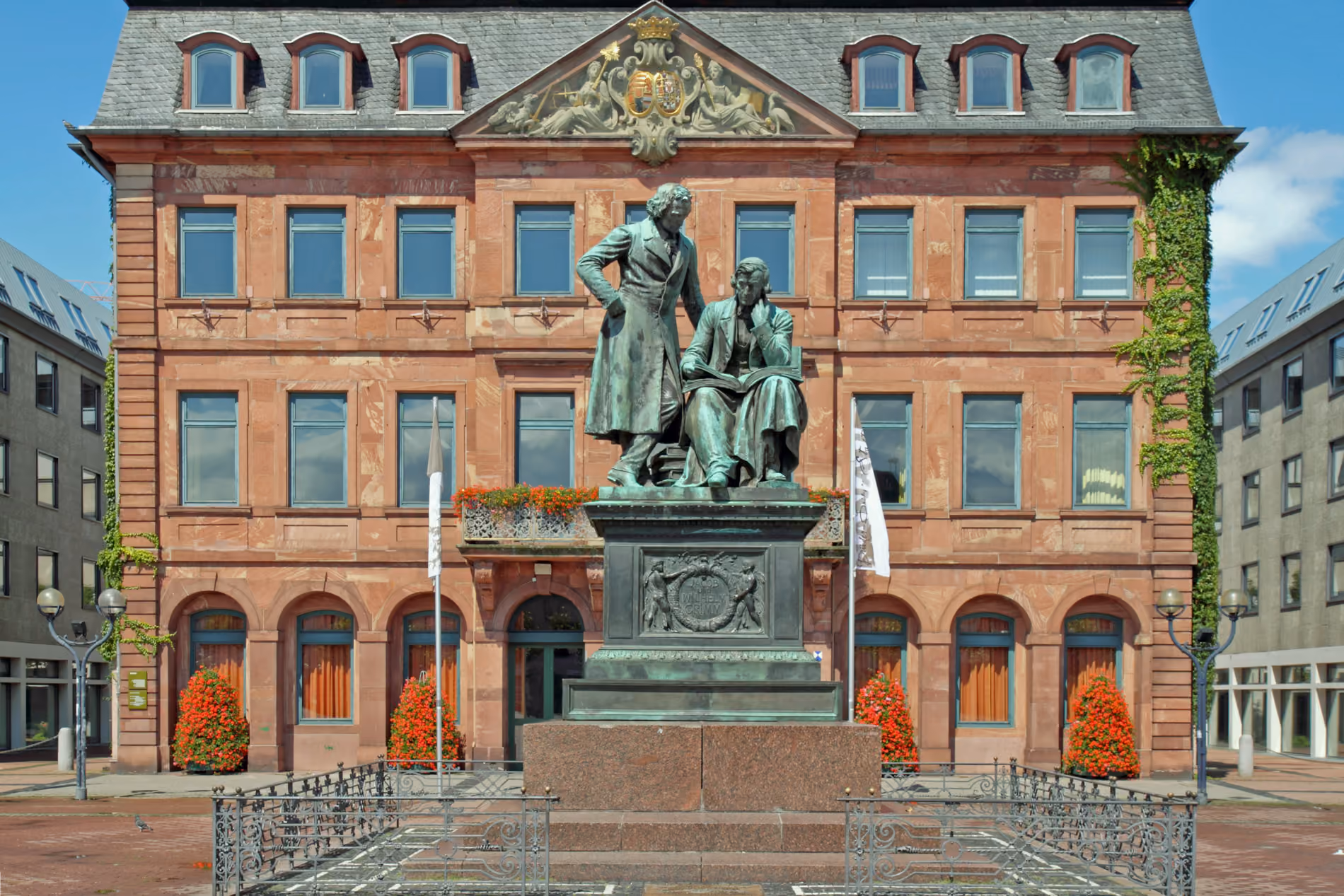 Bronzestatue der Brüder GrimmFiguren vor rotem Rathaus in Hanau mit Fenstern und Pflanzen