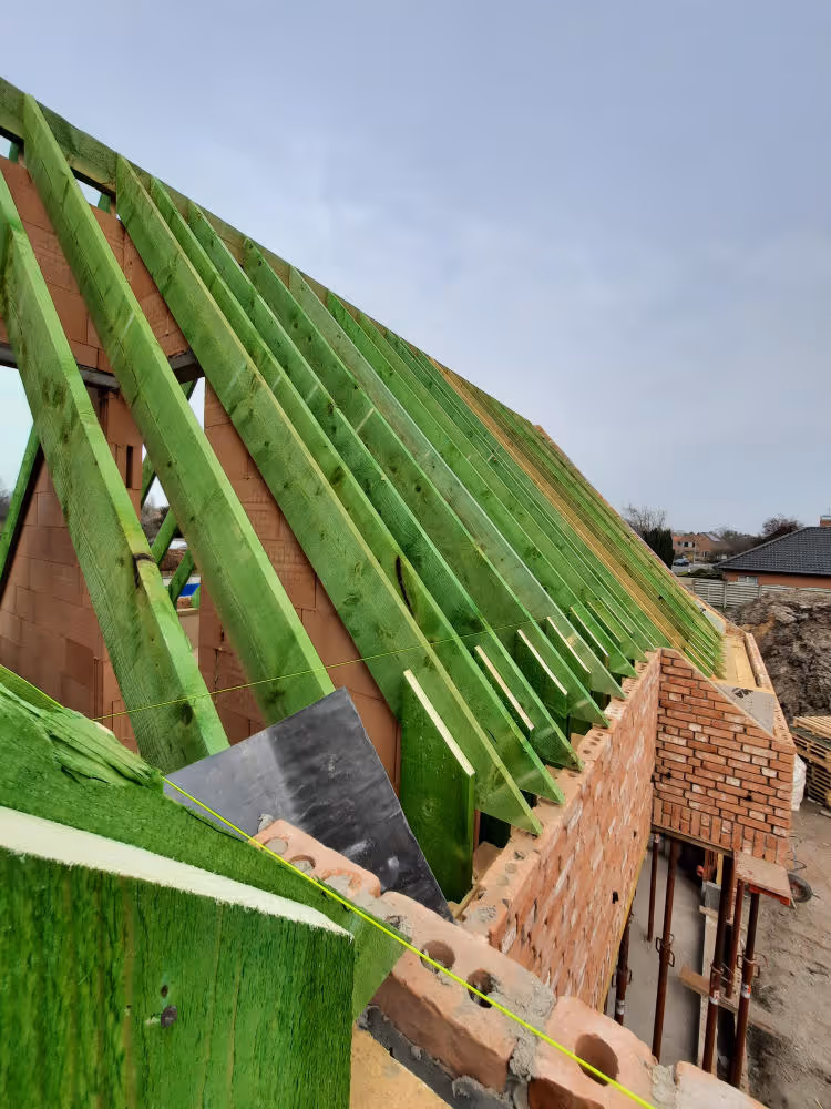 Green treated wooden roof beams installed on a brick house under construction.